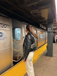 a woman in white pants and black jacket standing next to a silver train at a station