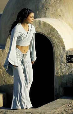 a woman standing in front of a stone wall wearing a white dress and shawl