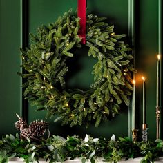 a wreath and candles on a mantel in front of a green wall with greenery