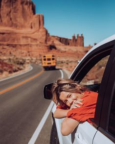 a woman leaning out the window of a car on a road with mountains in the background