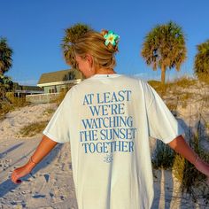 a woman standing on the beach with her hands in her pockets and wearing a t - shirt that says at least we're watching the sunset together