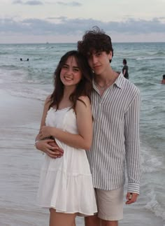 a young man and woman standing next to each other on the beach near the ocean