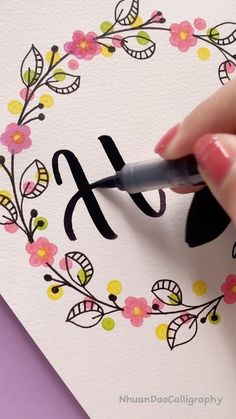 someone is writing the letter f on a card with flowers and leaves around it, while holding a marker