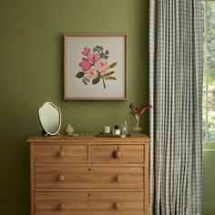 a dresser with flowers on it in front of a green wall and curtained window