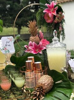 a table topped with pineapples and glasses filled with liquid next to flowers on top of a lush green field