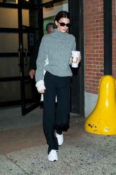 a woman in grey sweater and black pants walking down street with coffee cup on her hand