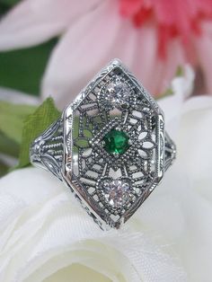 Simulated Green Emerald & White Cubic Zirconia (CZ) Sterling Silver RingCharlotte Design#D231 Resurrected from the Art Deco movement, this gorgeous Art Deco reproduction filigree ring is crafted in sterling silver. This lovely filigree ring is set with a stunning quality 2mm center round-cut simulated green emerald gemstone. Two small white cubic zirconia gemstones also grace the face of the ring, one near each point, each set in the center of a tiny heart. There are 2 filigree accent flowers th Heirloom Silver Filigree Ring With Intricate Design, Silver Emerald Ring With Intricate Design For Anniversary, Vintage Silver Filigree Ring With Diamond Accents, Anniversary Silver Emerald Ring With Intricate Design, Ornate Sterling Silver Filigree Ring With Intricate Design, Art Deco Silver Emerald Wedding Ring, Silver Art Deco Emerald Wedding Ring, Art Deco Sterling Silver Filigree Ring With Diamond Cut, Ornate Silver Diamond Ring With Filigree