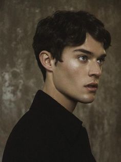 a young man with dark hair and blue eyes looks into the camera while standing in front of a wall