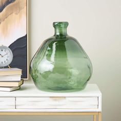 a green vase sitting on top of a white table next to a book and clock