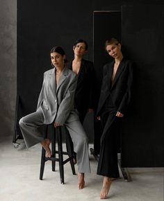 three models pose for the camera in front of a black wall, one sitting on a stool