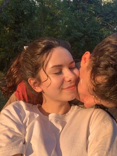 two women are kissing each other in front of some trees and bushes, one is wearing a white shirt