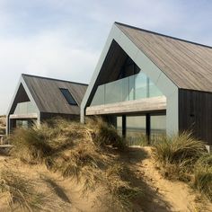 two houses on the beach with grass and sand dunes in front of them, one building is made of wood