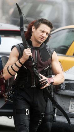 a man dressed in black holding a bow and arrow while walking down the street with cars behind him