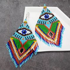 These XL Oversized Evil Eye Beaded Earrings feature a vibrant and intricate design, showcasing the iconic Evil Eye symbol in a unique and colorful palette. Crafted meticulously with a blend of light blue and green as the primary colors, the earrings also incorporate shades of blue, yellow, orange, black, and white. The bright and bold colors create a captivating look that stands out, while the long fringe adds a dynamic and playful element. Designed for those who appreciate bold and expressive j Evil Eye Symbol, Earrings Beads, Colorful Palette, Earrings Patterns, Bead Earring, Eye Symbol, Jaune Orange, Art Earrings, Earring Ideas