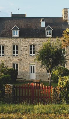 an old stone house with a red fence in front of it and trees around the yard