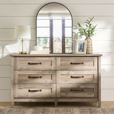 a dresser with a mirror on top of it next to a lamp and vase in front of a window