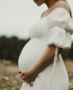 a pregnant woman standing in a field with her hands on her stomach, wearing a white dress