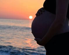 a pregnant woman standing in front of the ocean at sunset with her belly curled up