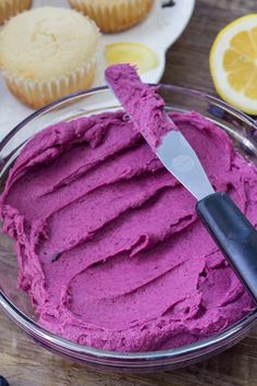 a bowl filled with purple frosting next to muffins and lemon wedges