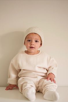 A young child sits against a plain, light-colored background, wearing a cozy cream-colored knit beanie and a matching soft, oversized sweatshirt and pants. The child looks forward with a calm expression. Knitted Baby Beanies, Baby Beanie Hat, Baby Beanie Hats, Silky Texture, Knitted Baby, Cozy Knit