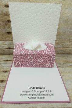 a pink and white card with a tissue box on it's side, in front of a wooden background