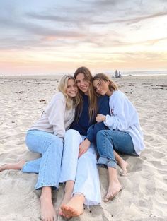 three women are sitting on the sand at the beach and one is holding her arm around another woman's shoulder