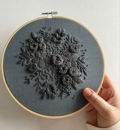 someone is holding up a hand embroidery project with black flowers on grey fabric and gold trimmings