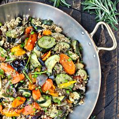 a pan filled with vegetables and rice on top of a wooden table next to a sprig of rosemary