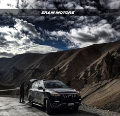 two men standing next to a parked car in the middle of mountains with clouds overhead