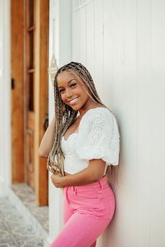 a woman leaning against a white wall wearing pink pants and a white top with braids