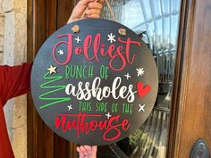 Introducing the Jolliest Bunch of Assholes this Side of the Nuthouse door sign – a whimsical and humorous addition to your festive décor! Stand out with the perfect blend of humor and holiday spirit in your home. A B O U T • T H I S • I T E M✔ Measures roughly 17.5" in diameter.✔ Constructed of 1/4" premium Baltic birch ply.✔ Stylish black backboard for a sleek and modern look.✔ Vibrant red, green, and white painted cheery fonts for a festive touch.✔ Laser-cut letters and designs are adhered as Cute Christmas Signs Diy, Jolliest Bunch This Side Of The Nuthouse, Funny Christmas Door Signs, Christmas Door Signs, Cricut Clothes, Holiday Wood Sign, Laser Crafts, Christmas Signs Diy, Hanger Ideas