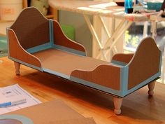 a doll bed sitting on top of a wooden floor next to a table with paper and scissors