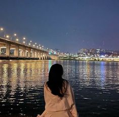 a woman is sitting on the shore looking at the water and bridge in the background