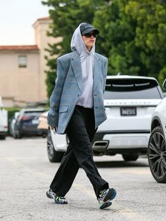 a woman walking across a parking lot wearing a gray jacket and black pants with a hoodie over her head