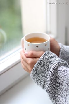 a woman holding a cup of tea in front of a window