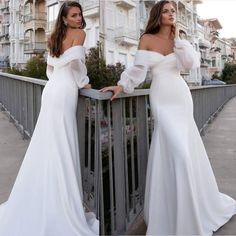 two beautiful women in white dresses standing on a bridge with their arms around each other