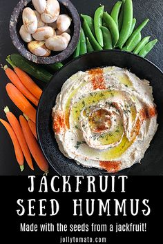 a bowl of hummus next to carrots and garlic on a black plate with the title jackfrut seed hummus made with the seeds from a jackfruit