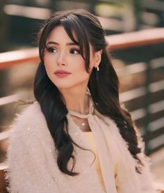 a woman with long black hair sitting on a bench wearing a white coat and pearl earrings