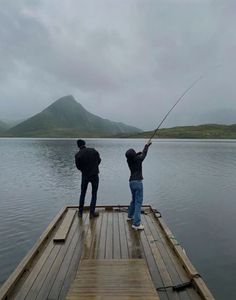 two people standing on a dock fishing in the water with mountains in the background and cloudy skies