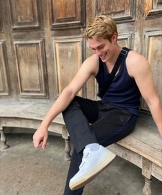 a young man is sitting on a wooden bench and smiling at the camera while wearing white sneakers