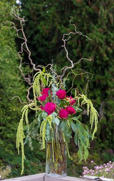 a vase filled with pink roses and greenery on top of a wooden table in front of trees