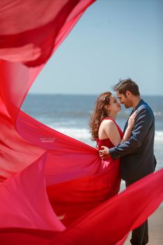 a man and woman standing next to each other near the ocean with red fabric blowing in the wind