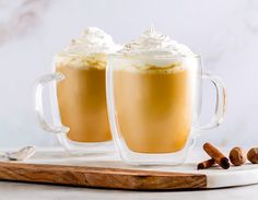 two mugs filled with hot chocolate and whipped cream on a cutting board next to cinnamon sticks