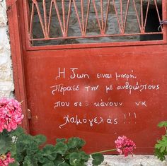 a red door with writing on it next to some pink flowers and green plants in front of it