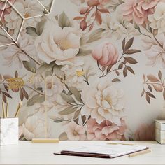 a desk with a book, notebook and pen in front of a floral wallpaper