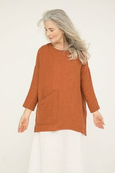 an older woman with grey hair wearing a brown top and white skirt, standing in front of a white background