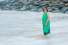 a woman in a green dress standing in the water at the beach with rocks behind her