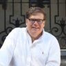 a man with glasses and a white shirt is sitting down in front of a building