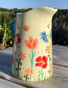 a painted pitcher sitting on top of a wooden table next to grass and flowers in the background