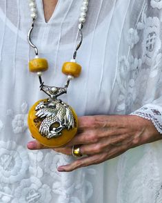 The majesty of the Himalayas. Tibetan yellow copal pendant embossed with Tibetan silver images of the Phoenix and the Elephant. Hill Tribe pure silver (.999) spacers, white fresh water pearls, vintage Tibetan copal beads, sterling silver beads, sterling silver clasp.  Length: 29" Pendant: 4"  Powerful and unique, this 29" long necklace centers on a large yellow Tibetan copal pendant, which is embossed with Tibetan "silver". A two-sided pendant, one side depicts the Elephant, which represents strength of mind in Tibetan Buddhism, the other side depicts the Phoenix, which symbolizes tranquility and peace. I have used vintage yellow Tibetan copal beads, large white pearls, and large spacers made of Hill Tribe pure silver (.999). It is definitely a powerful necklace that exudes the vast spirit Yellow Pendant Jewelry For Festivals, Carved White Gold Pendant Jewelry, Carved Silver Medallion Jewelry, Carved Amulet Jewelry For Collectors, Ceremonial Carved Amulet Jewelry, Silver Carved Necklaces, Traditional Carved Round Pendant Jewelry, Silver Carved Round Necklace, Artisan Carved Amber Necklace